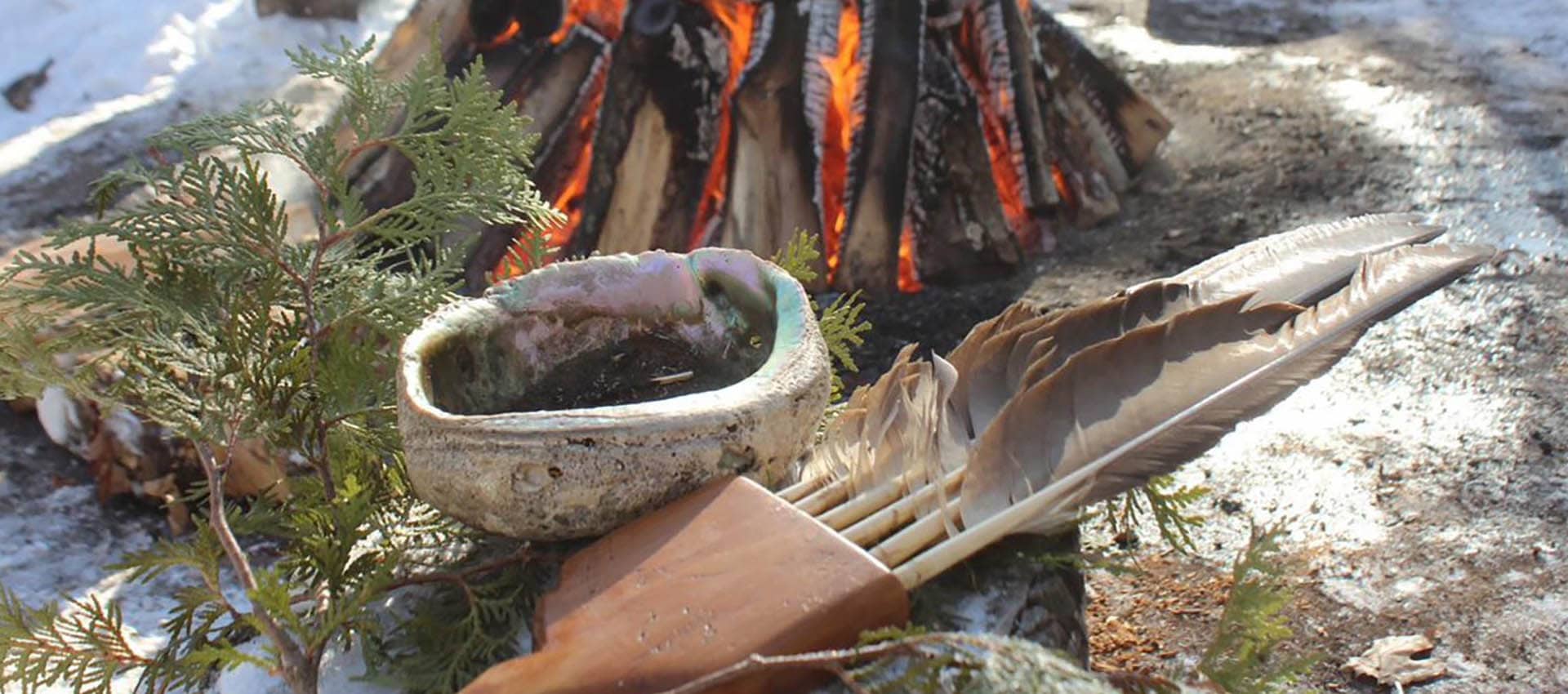 Cedar branches, abalone smudge bowl and feather fan sitting in front of a fire